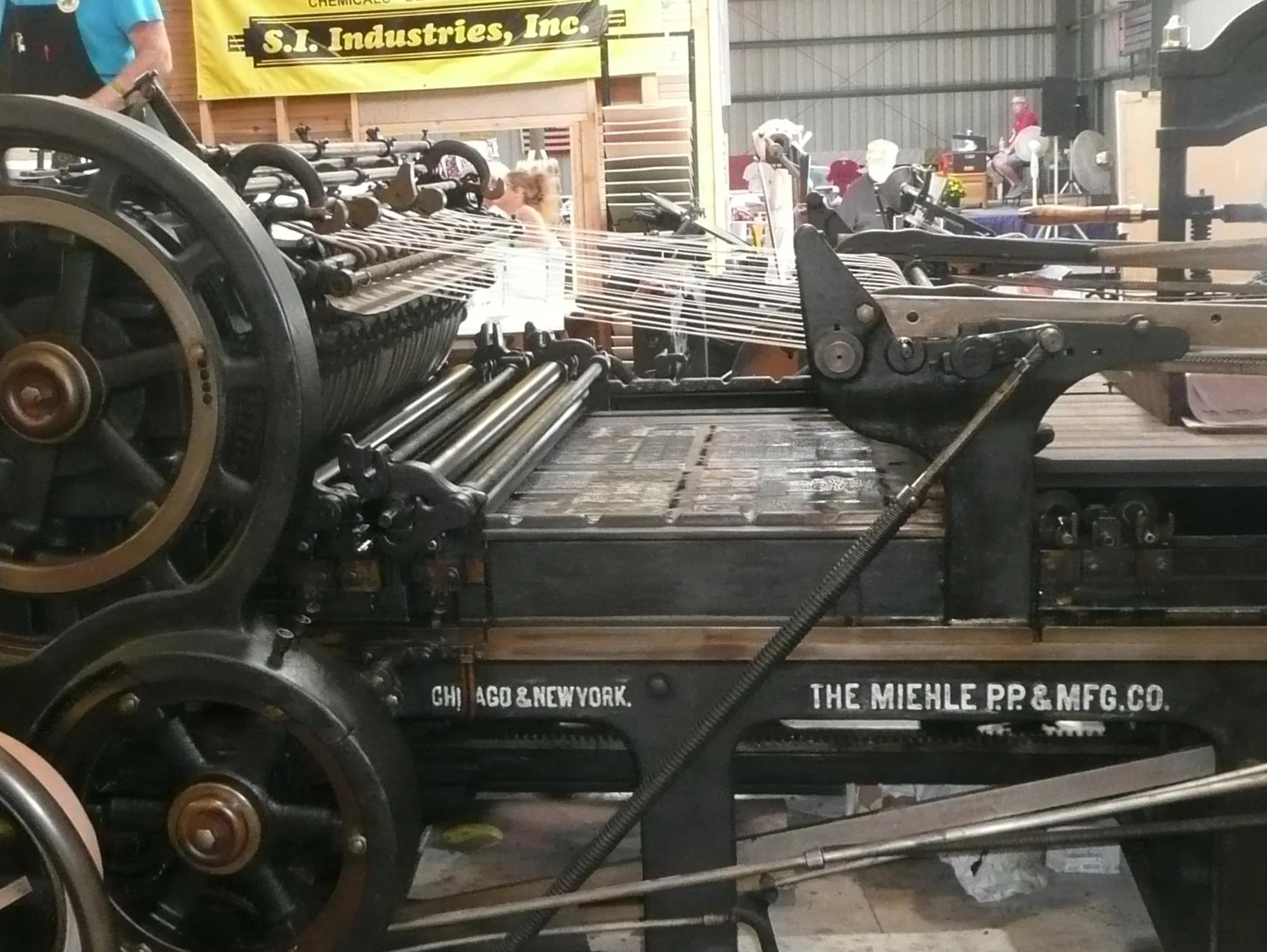 The bed of metal type passing under the inking rollers of a Miehle Model 00 flat-bed cylinder printing press . The print roller is on the left; the press operator, in the upper left, loads pages to be printed, while the webbing above the bed delivers printed pages to be lifted onto the output tray o. Print shop expansion: four routes and when each fits
